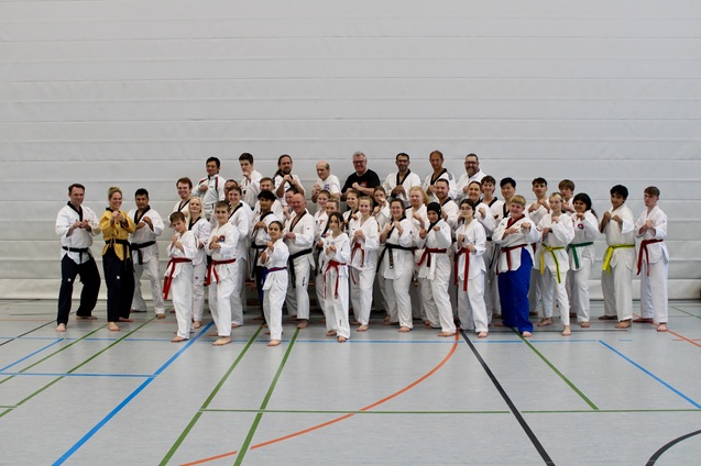 Gruppenshot von Karateka in weißen Anzügen, einige tragen farbige Gürtel, auf einem Sportplatz in einer Halle.
