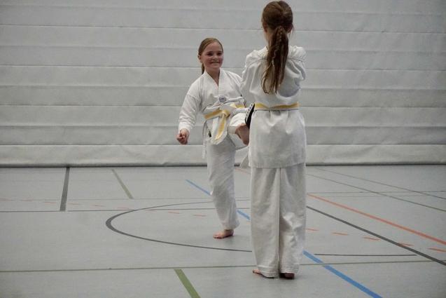 Zwei Kinder in weißen Karateanzügen üben gegenseitig einen Kick auf einer Sportplatzfläche.