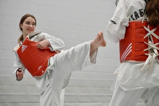 Mädchen in weißem Taekwondo-Anzug führt High Kick gegen einen Trainingspartner in rotem Schutzanzug aus.