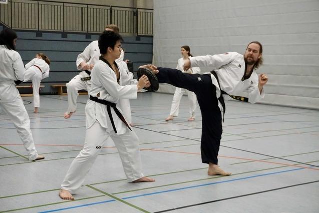 Taekwondo-Training in einer Sporthalle mit Kämpfern in weißen Uniformen. Ein Kämpfer führt einen Kick aus.