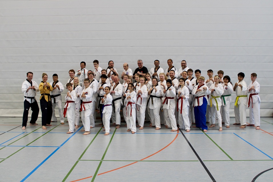 Gruppenshot von Karateka in wei&szlig;en Anz&uuml;gen, einige tragen farbige G&uuml;rtel, auf einem Sportplatz in einer Halle.