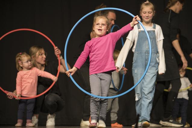 Mädchen in rosa Oberteil hält einen blauen Hula-Hoop-Reifen, während andere Kinder mit Reifen auf der Bühne stehen.