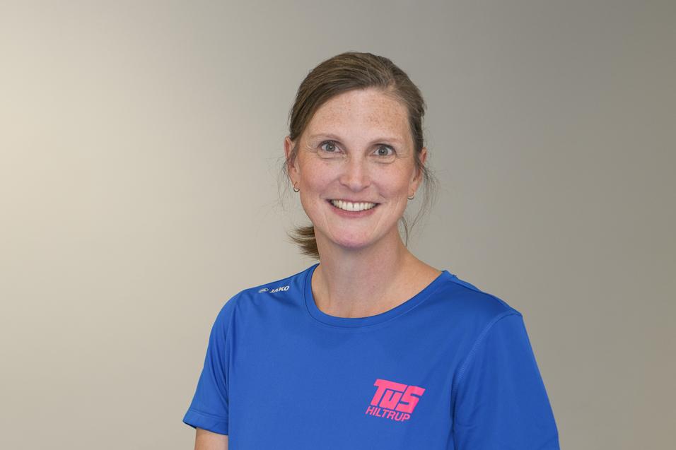 Lächelnde Frau in blauem Sportshirt mit pinkem Logo, vor neutralem Hintergrund.