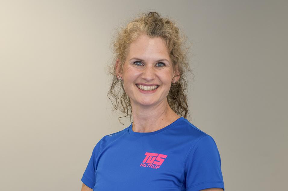 Porträt einer lächelnden Frau mit lockigem Haar in einem blauen Shirt vor einem neutralen Hintergrund.