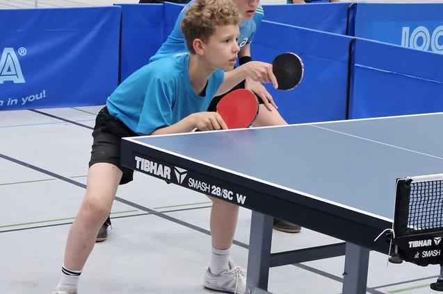 Zwei Jungen in blauen T-Shirts stehen an einem Tischtennistisch, bereit f&uuml;r einen Schlag mit Schl&auml;gern in der Hand.