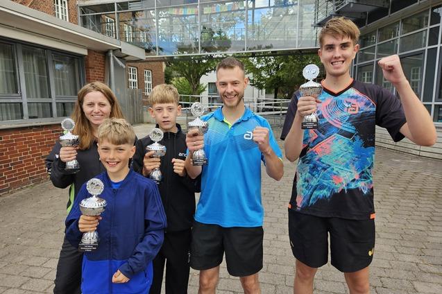 Fünf Personen halten Pokale vor einem Gebäude, alle zeigen freudig ihre Auszeichnungen und posieren für das Foto.