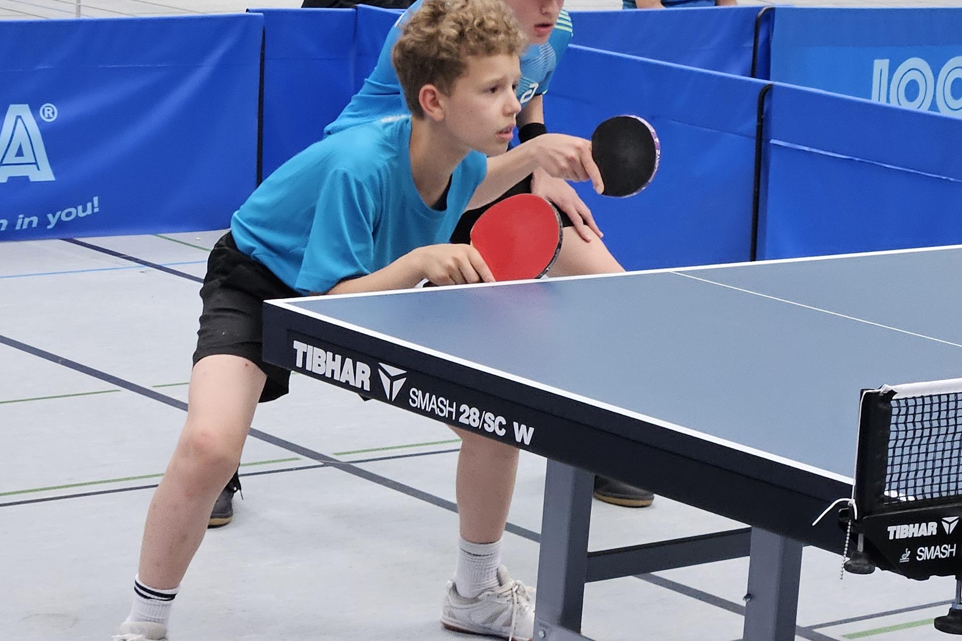 Zwei Jungen in blauen T-Shirts stehen an einem Tischtennistisch, bereit für einen Schlag mit Schlägern in der Hand.