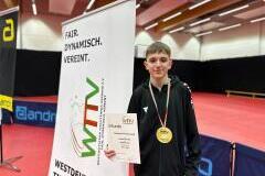 Jugendlicher mit Goldmedaille und Urkunde steht neben einem WTTV-Banner in einer Tischtennishalle.