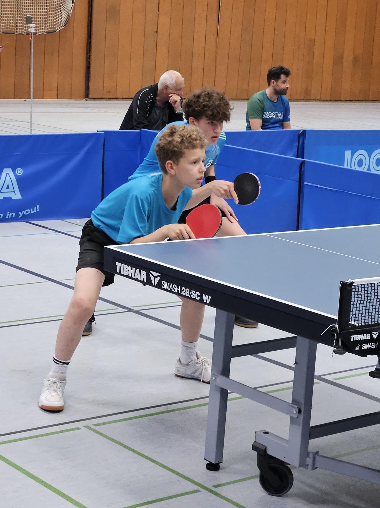 Zwei Jungen in blauen T-Shirts stehen an einem Tischtennistisch, bereit für einen Schlag mit Schlägern in der Hand.