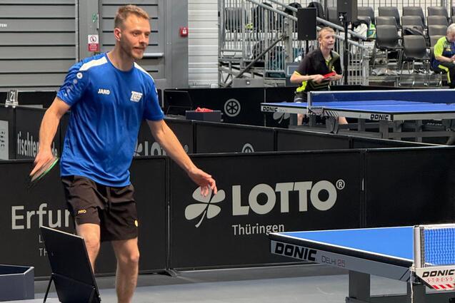 Mann im blauen Trikot spielt Tischtennis, während ein anderer Spieler im Hintergrund sich vorbereitet. Tischtennisplatte sichtbar.