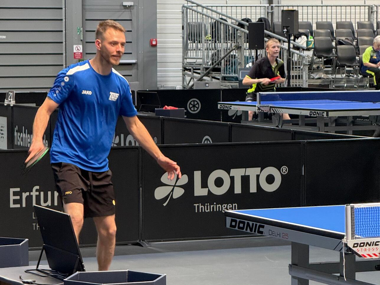 Mann im blauen Trikot spielt Tischtennis, während ein anderer Spieler im Hintergrund sich vorbereitet. Tischtennisplatte sichtbar.