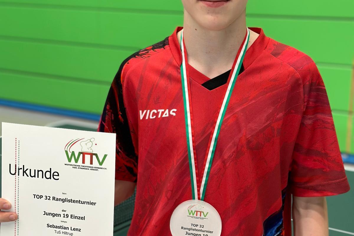 Teenager mit Medaille und Urkunde für den 1. Platz im Tischtennisturnier, hinter ihm eine grüne Tischtennisplatte.