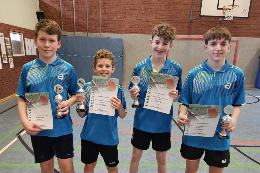 Vier Jungen in blauen Trikots halten Pokale und Urkunden in einer Sporthalle mit Tischtennispl&auml;tzen.