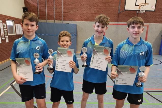 Vier Jungen in blauen Trikots halten Pokale und Urkunden in einer Sporthalle mit Tischtennispl&auml;tzen.