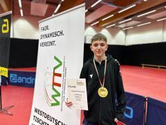Jugendlicher mit Goldmedaille und Urkunde steht neben einem WTTV-Banner in einer Tischtennishalle.