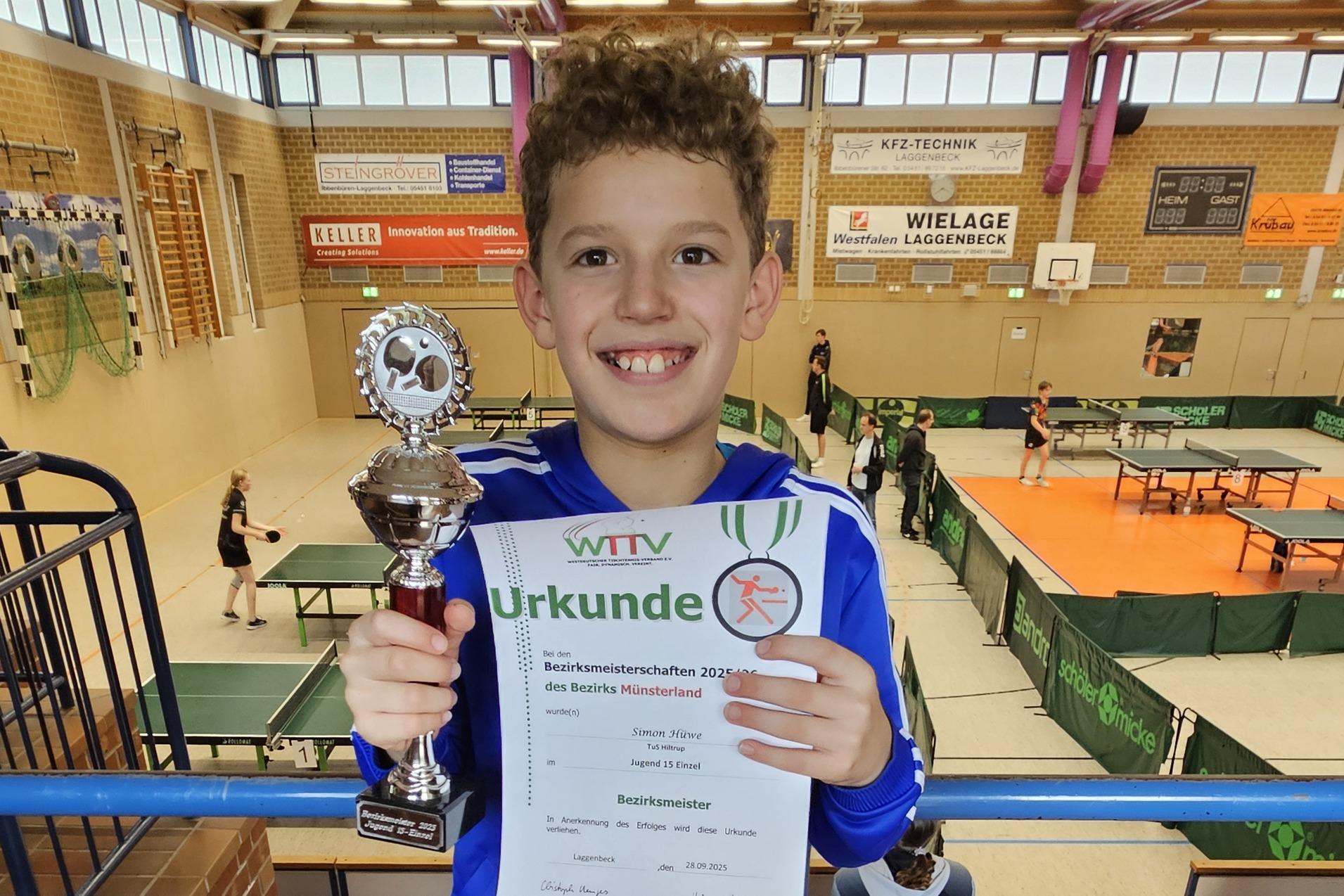 Junge in blauer Jacke h&auml;lt Pokal und Urkunde nach Tischtennisturnier in Sporthalle mit Tischen im Hintergrund.