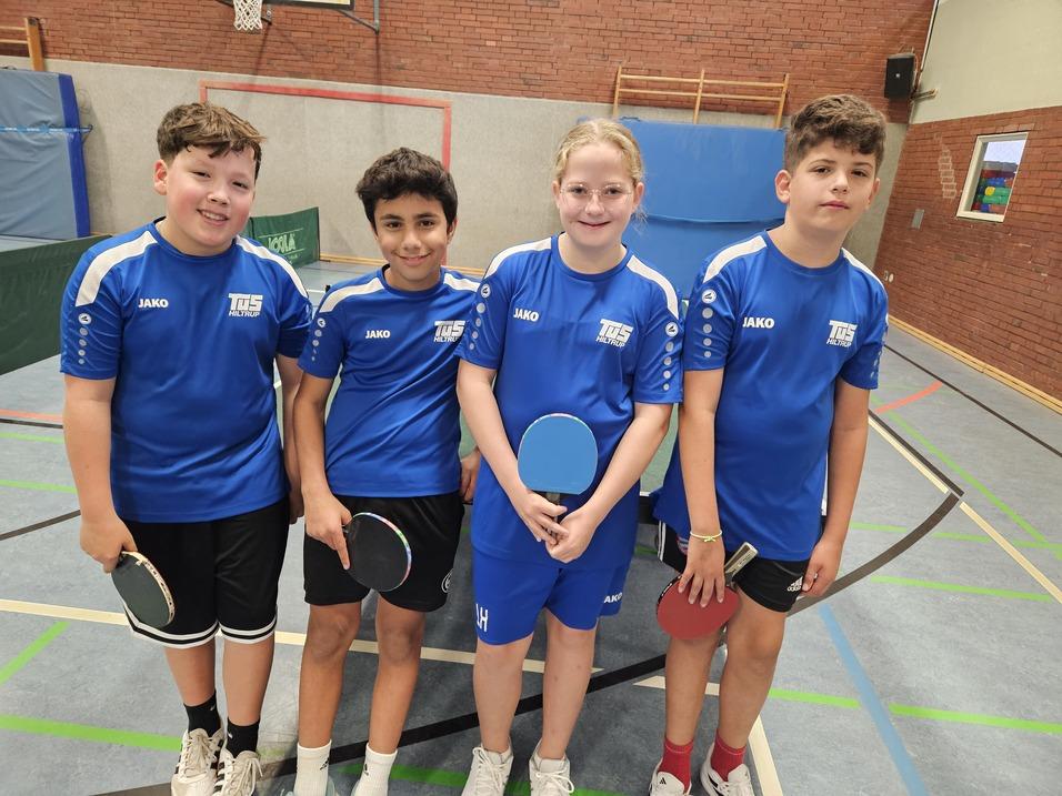 Vier Kinder in blauen Sporttrikots stehen lächelnd nebeneinander, jeder mit einem Tischtennis-Schläger in der Hand.