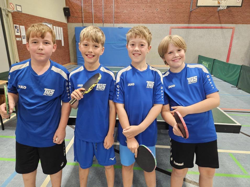 Vier Kinder in blauen Tischtennis-Trikots stehen lächelnd vor einem Tischtennistisch, jeder mit einem Schläger in der Hand.