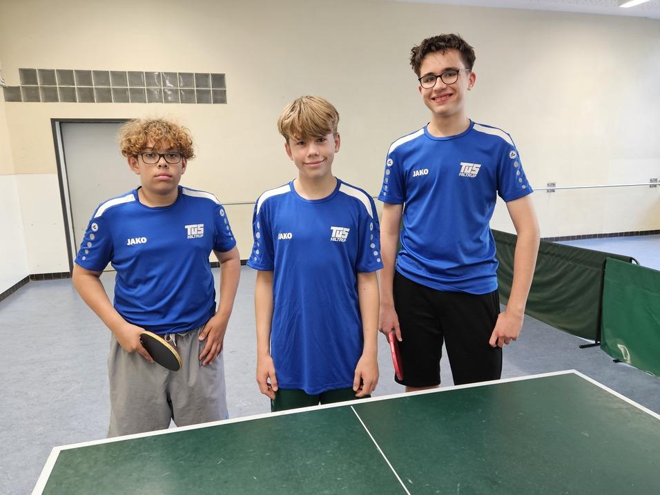 Drei Jungen in blauen Tischtennistrikots stehen neben einem Tischtennistisch in einer Hallenansicht.