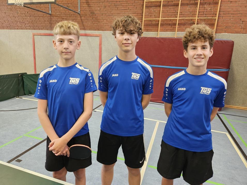 Drei Jugendliche in blauen Trikots stehen auf einem Tischtennisplatz, bereit für ein Match mit Schläger in der Hand.