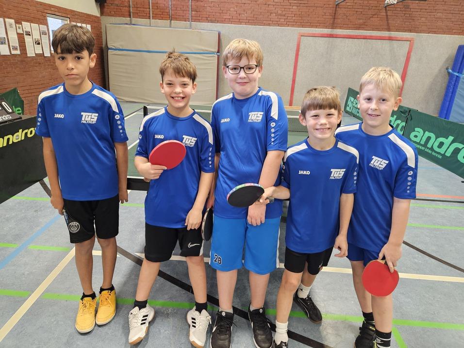 Fünf Kinder in blauen Tischtennis-T-Shirts stehen lächelnd mit Schlägern und Bällen in einer Sporthalle.