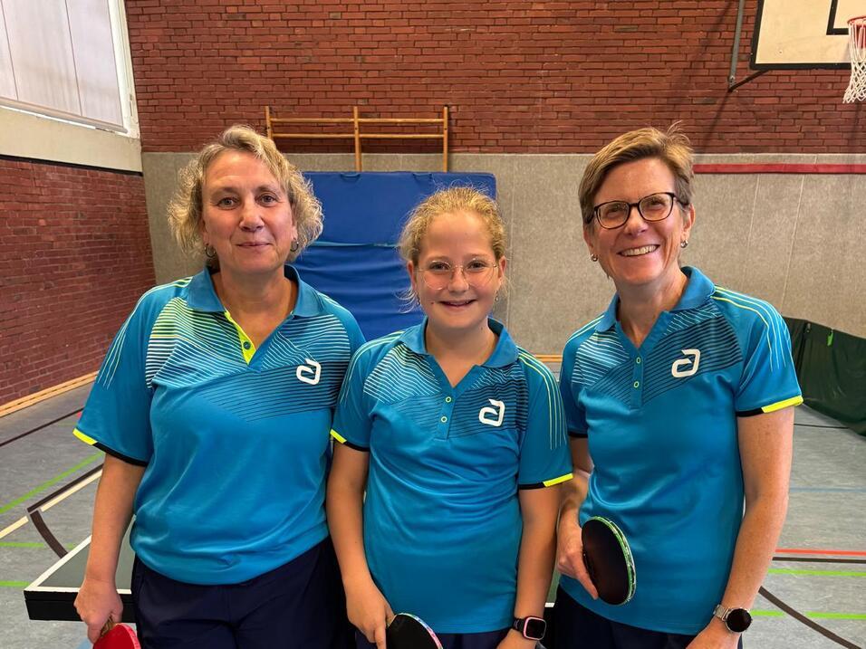 Drei Tischtennisspielerinnen, eine Erwachsene und ein Kind, posieren in blauen Trikots mit Schlägern in einer Sporthalle.