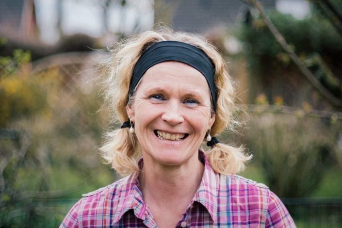 L&auml;chelnde Frau mit blonden Locken und Haarband, tr&auml;gt ein kariertes Hemd, steht drau&szlig;en vor gr&uuml;ner Landschaft.