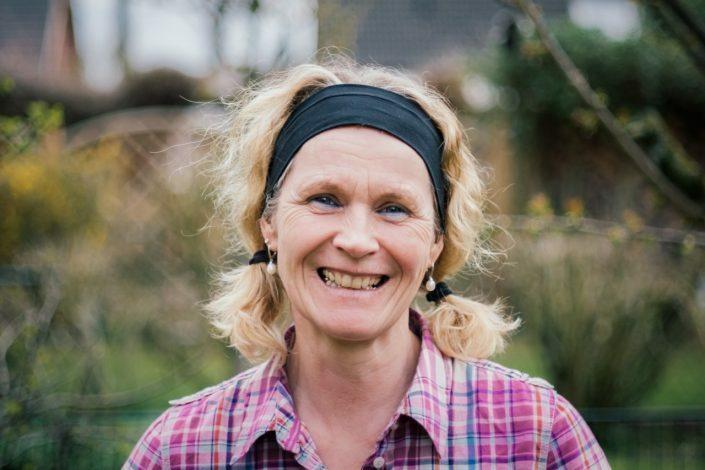 Lächelnde Frau mit blonden Locken und Haarband, trägt ein kariertes Hemd, steht draußen vor grüner Landschaft.