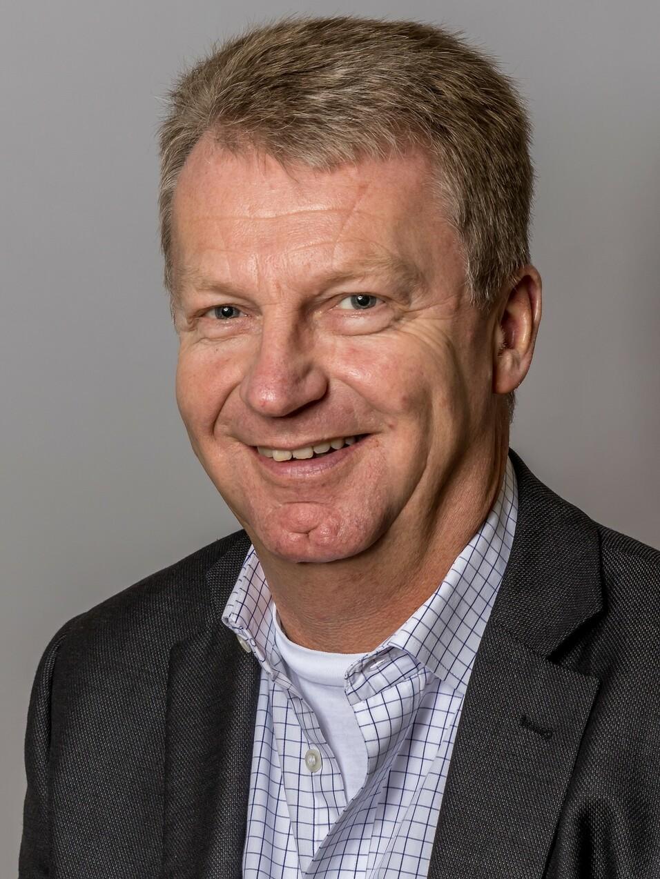 Porträt eines lächelnden Mannes mit kurzen, hellen Haaren, in einem schwarzen Blazer und karierten Hemd. Heller Hintergrund.