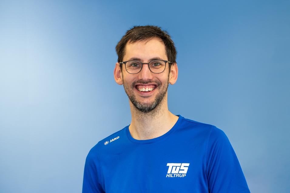 L&auml;chelnder Mann mit Brille tr&auml;gt ein blaues Sport-T-Shirt vor einem hellblauen Hintergrund.