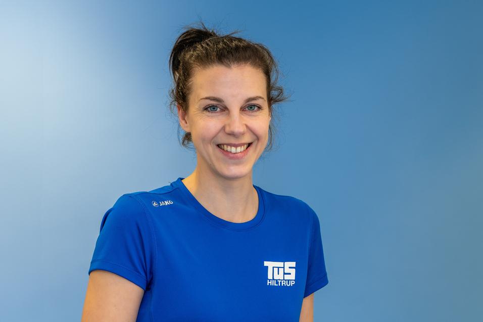 L&auml;chelnde Frau in blauer Sportbekleidung vor einem hellblauen Hintergrund, tr&auml;gt das Logo von TuS Hiltrup.