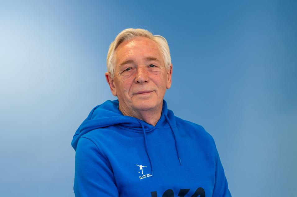 &Auml;lterer Mann mit kurzen grauen Haaren, tr&auml;gt einen blauen Kapuzenpullover, vor einfarbig blauer Wand.