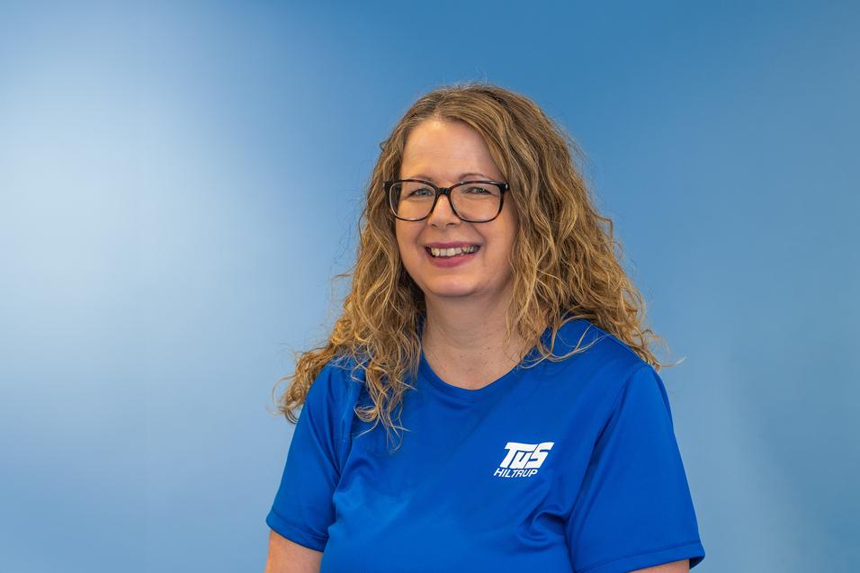 Frau mit lockigem, blondem Haar und Brille, l&auml;chelnd in blauem T-Shirt vor einem hellblauen Hintergrund.