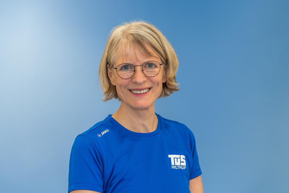 Porträt einer lächelnden Frau mit blonden, schulterlangen Haaren in einem blauen Sportshirt vor einem hellblauen Hintergrund.