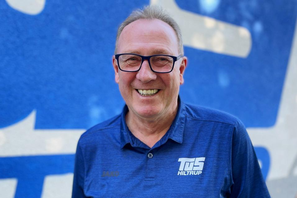 Portr&auml;t eines l&auml;chelnden Mannes mit Brille, in einem blauen Shirt mit dem Logo "TuS Hiltrup" vor einer blauen Wand.