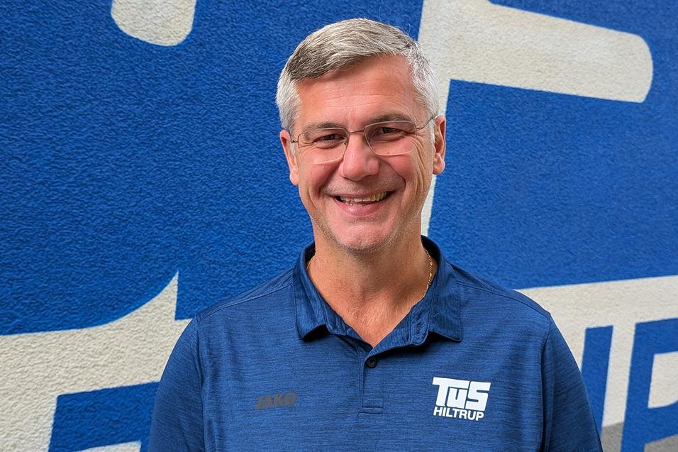 L&auml;chelnder Mann mit Brille in einem blauen Sportshirt vor einer blauen Wand mit wei&szlig;en Grafiken.