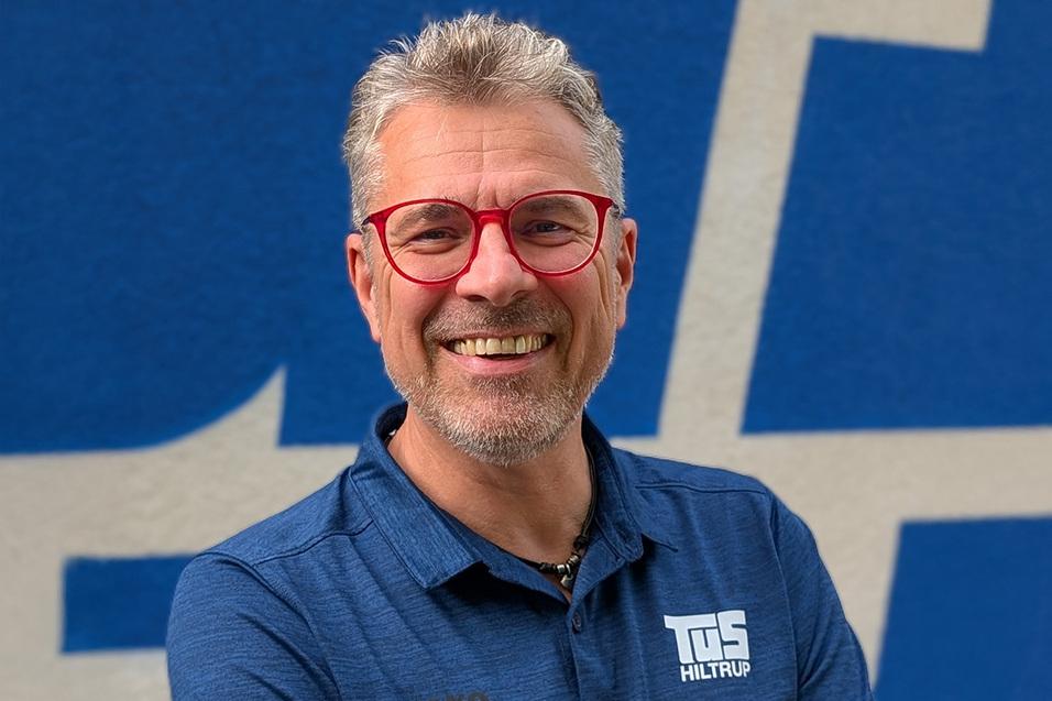 L&auml;chelnder Mann mit grauen Haaren und roten Brillenfassungen, tr&auml;gt ein dunkelblaues Shirt vor einem blauen Hintergrund.