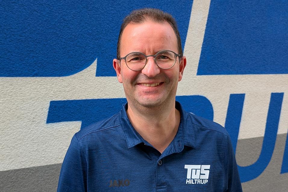 Andree Goldbeck Mann mit Brille und lächelndem Gesicht trägt ein blaues Hemd vor einer graublauen Wand mit Schriftzug.