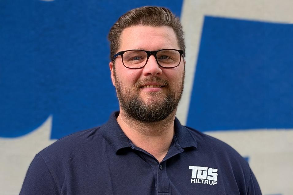 Thomas Rohoff Portrait eines Mannes mit Brille und Bart, trägt ein dunkles Poloshirt mit dem Logo von TuS Hiltrup, vor blauem Hintergrund.