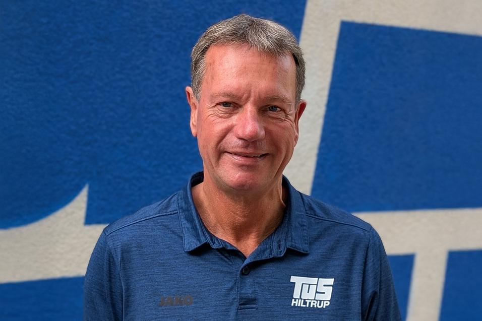 Uli Dütting Mann steht vor einer blauen Wand, trägt ein dunkles Polohemd mit dem Logo von TuS Hiltrup.