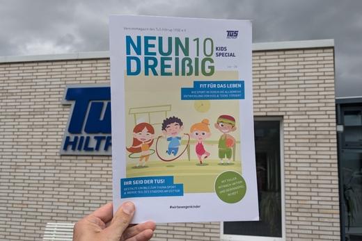 Hand h&auml;lt ein Vereinsmagazin des TuS Hiltrup mit dem Titel &bdquo;NEUN DREI&szlig;IG KIDS SPECIAL&ldquo; vor einer Geb&auml;ude-Fassade.
