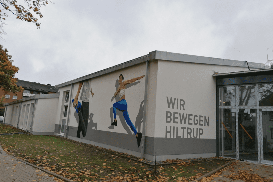 Wandmalerei an einem Gebäude zeigt zwei Sportler in dynamischen Posen mit dem Schriftzug „WIR BEWEGEN HILTRUP“.