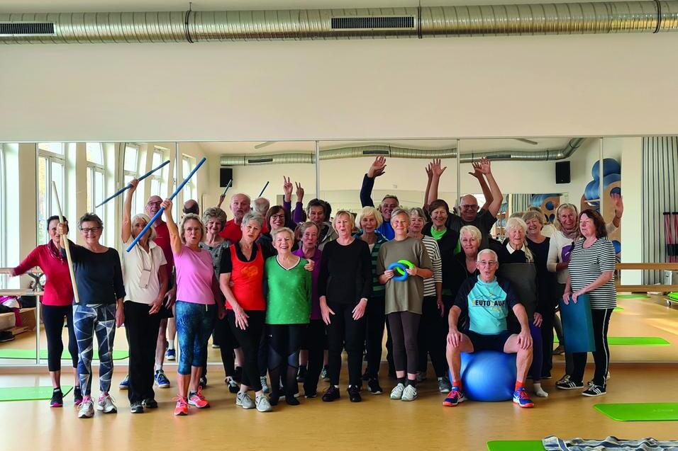 Gruppe älterer Menschen in Sportkleidung posiert mit Fitnessgeräten in einem Kursraum vor einem großen Spiegel.