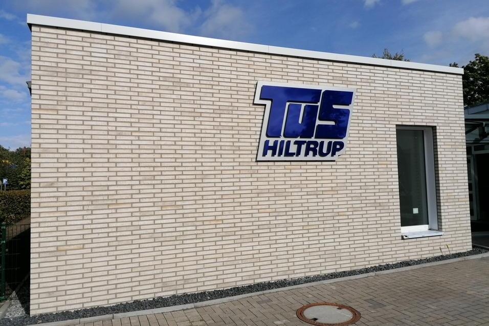 Fassade eines Gebäudes mit dem blauen Schriftzug „TuS Hiltrup“ auf hellen Ziegeln und einem Fenster.