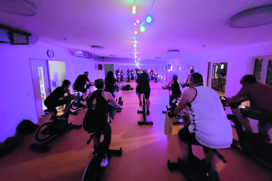 Gruppentraining beim Indoor-Cycling in einem dunkel beleuchteten Raum mit bunten Lichtern und Teilnehmern auf Uniformen.