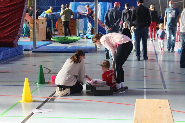 Zwei Erwachsene sitzen mit einem Kind auf dem Boden in einer Turnhalle, umgeben von aktiv spielenden Kindern und Spielgeräten.