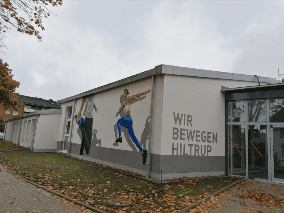 Wandmalerei an einem Gebäude zeigt zwei Sportler in dynamischen Posen mit dem Schriftzug „WIR BEWEGEN HILTRUP“.