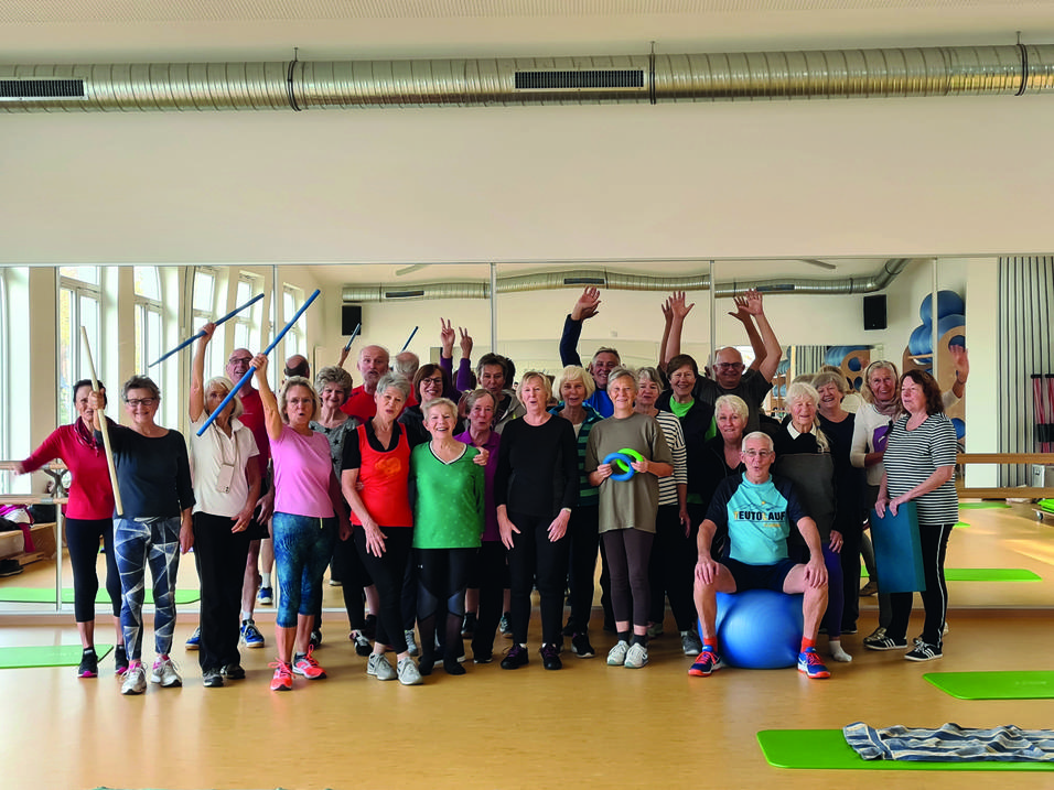 Gruppe älterer Menschen in Sportkleidung posiert mit Fitnessgeräten in einem Kursraum vor einem großen Spiegel.