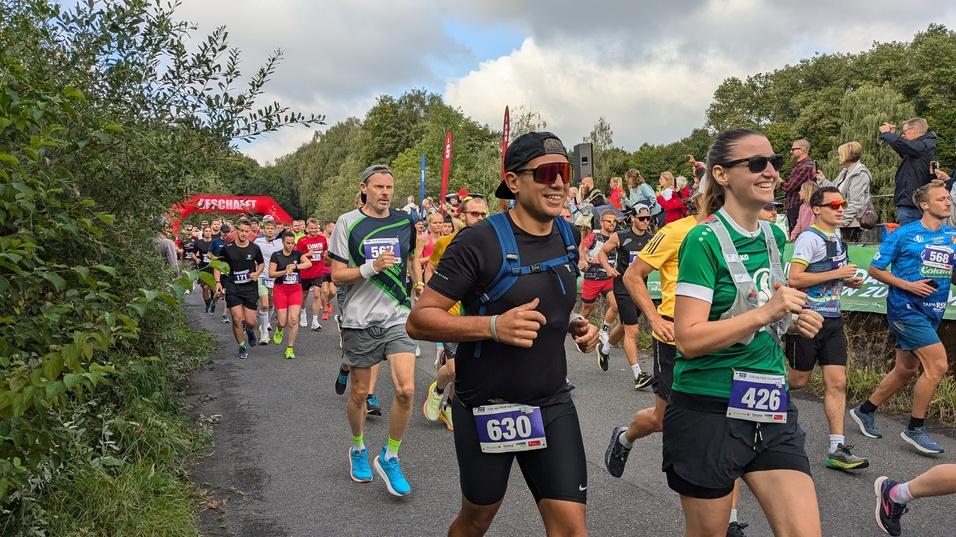 Gruppe von Läufern beim Wettkampf auf einer Straße, mit Zuschauern im Hintergrund und einem Laufbogen in der Ferne.