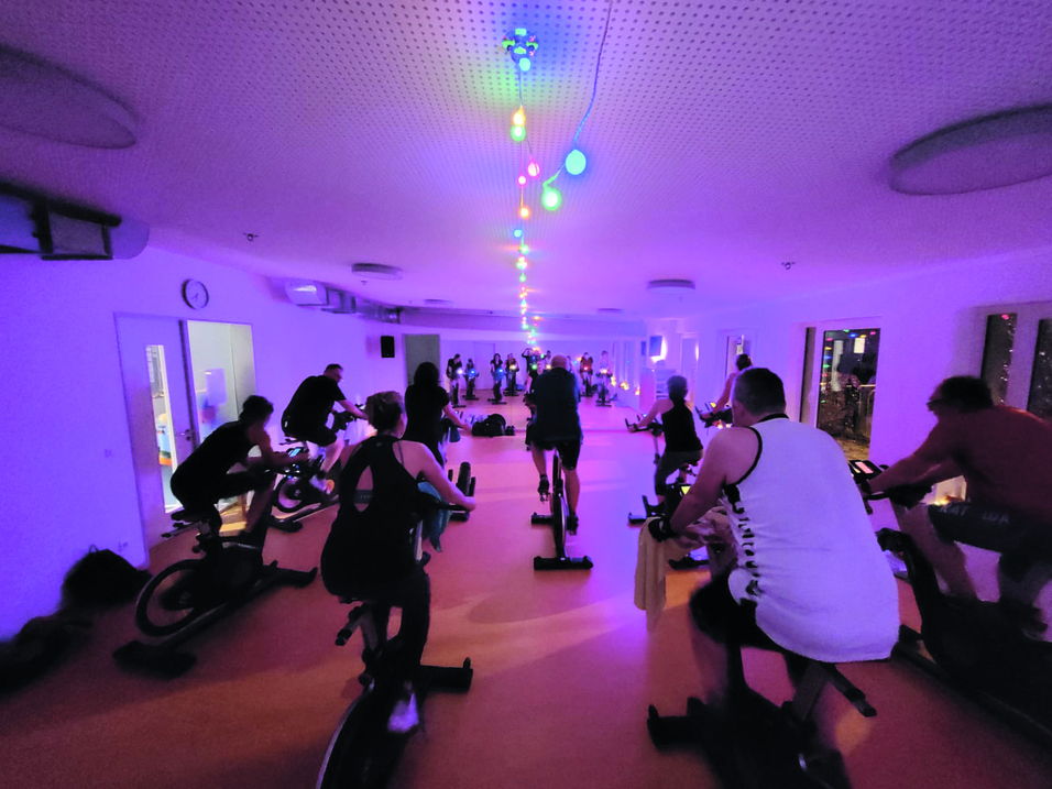 Gruppentraining beim Indoor-Cycling in einem dunkel beleuchteten Raum mit bunten Lichtern und Teilnehmern auf Uniformen.
